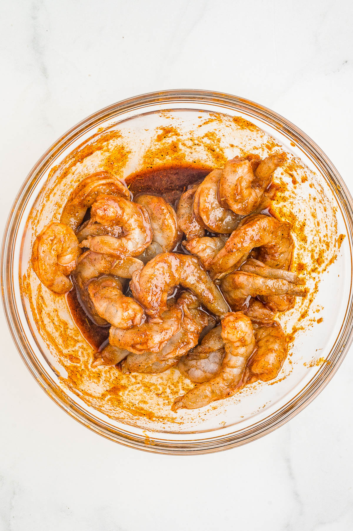Raw shrimp coated in a reddish marinade sitting in a clear glass bowl on a white surface.