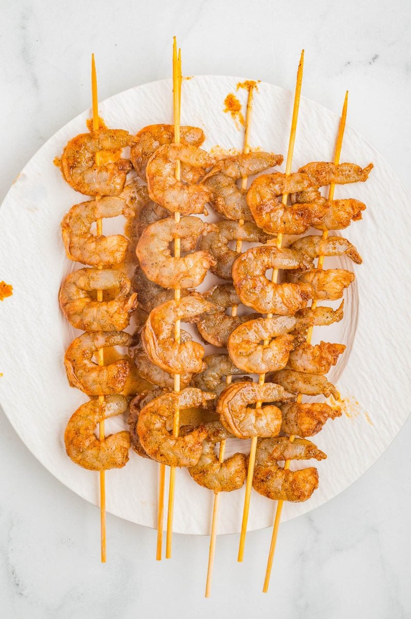 Five skewers of grilled, seasoned shrimp arranged on a white plate, viewed from above.