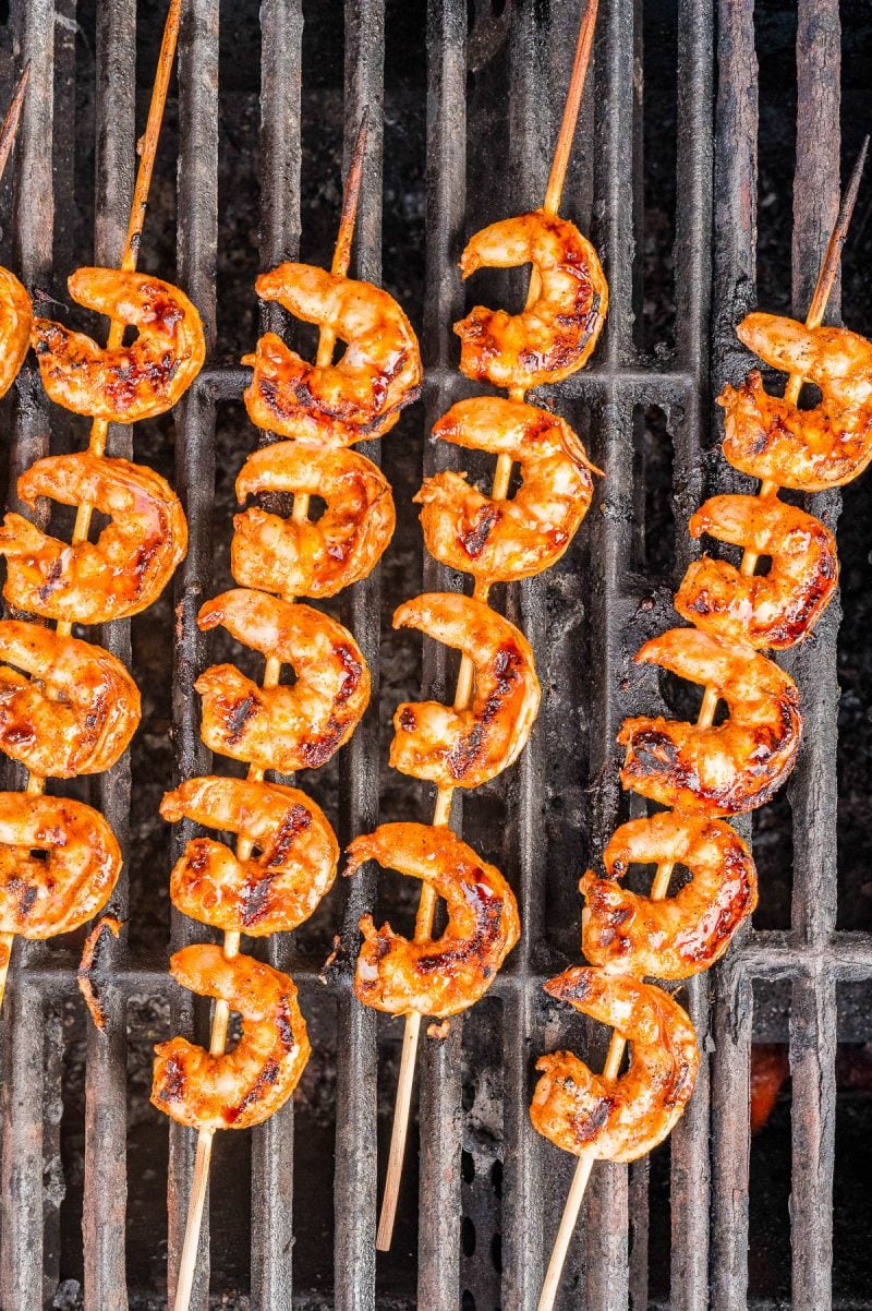 Four wooden skewers with grilled shrimp cooking on a barbecue grill, showing char marks and a reddish seasoning.
