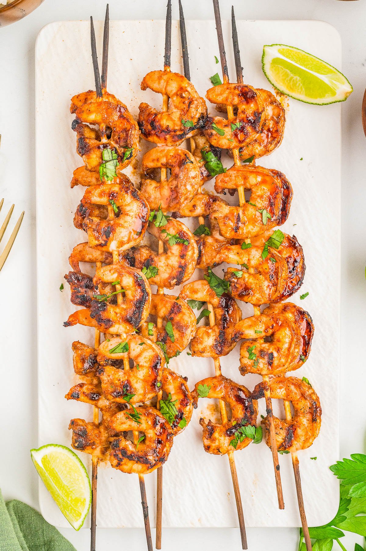 Four skewers of grilled shrimp garnished with chopped herbs, served on a white tray with lime wedges on the side.