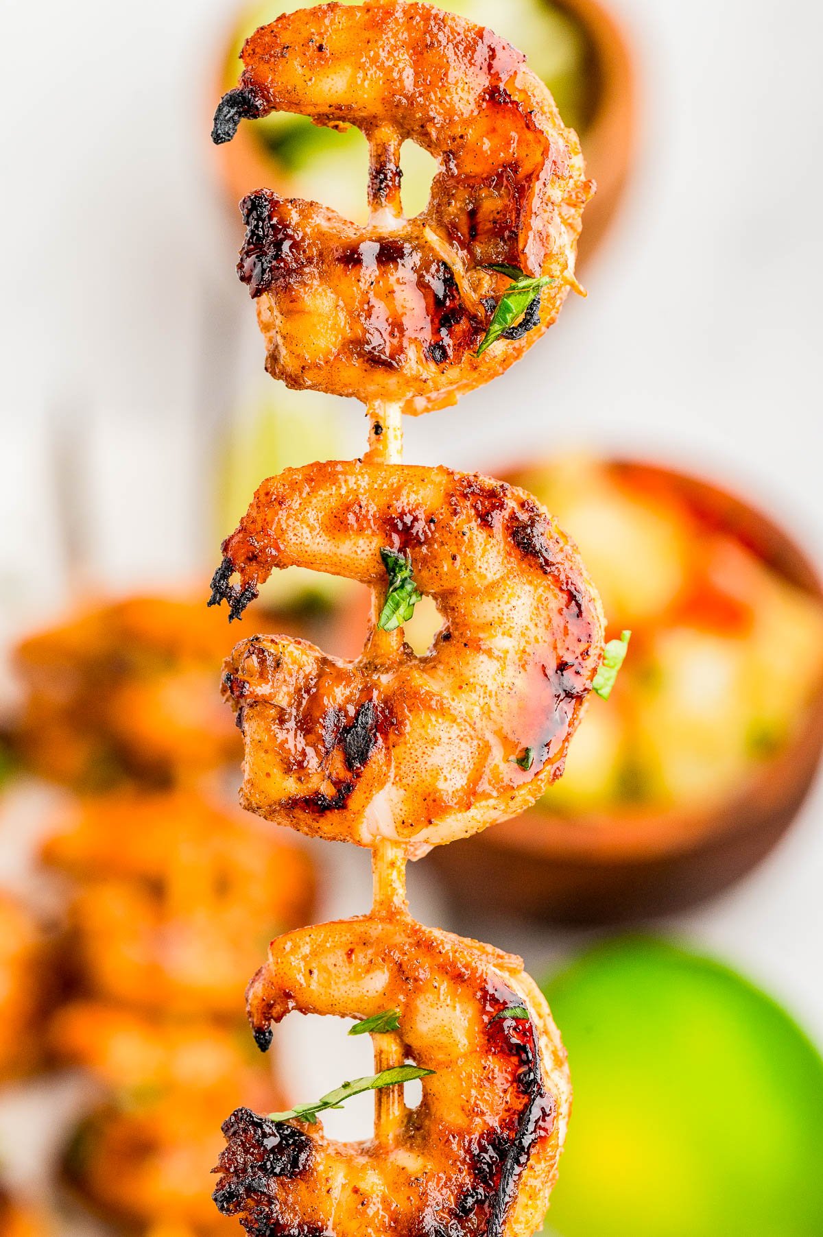 Close-up of grilled shrimp skewers, seasoned and slightly charred, with a blurred background featuring various foods.