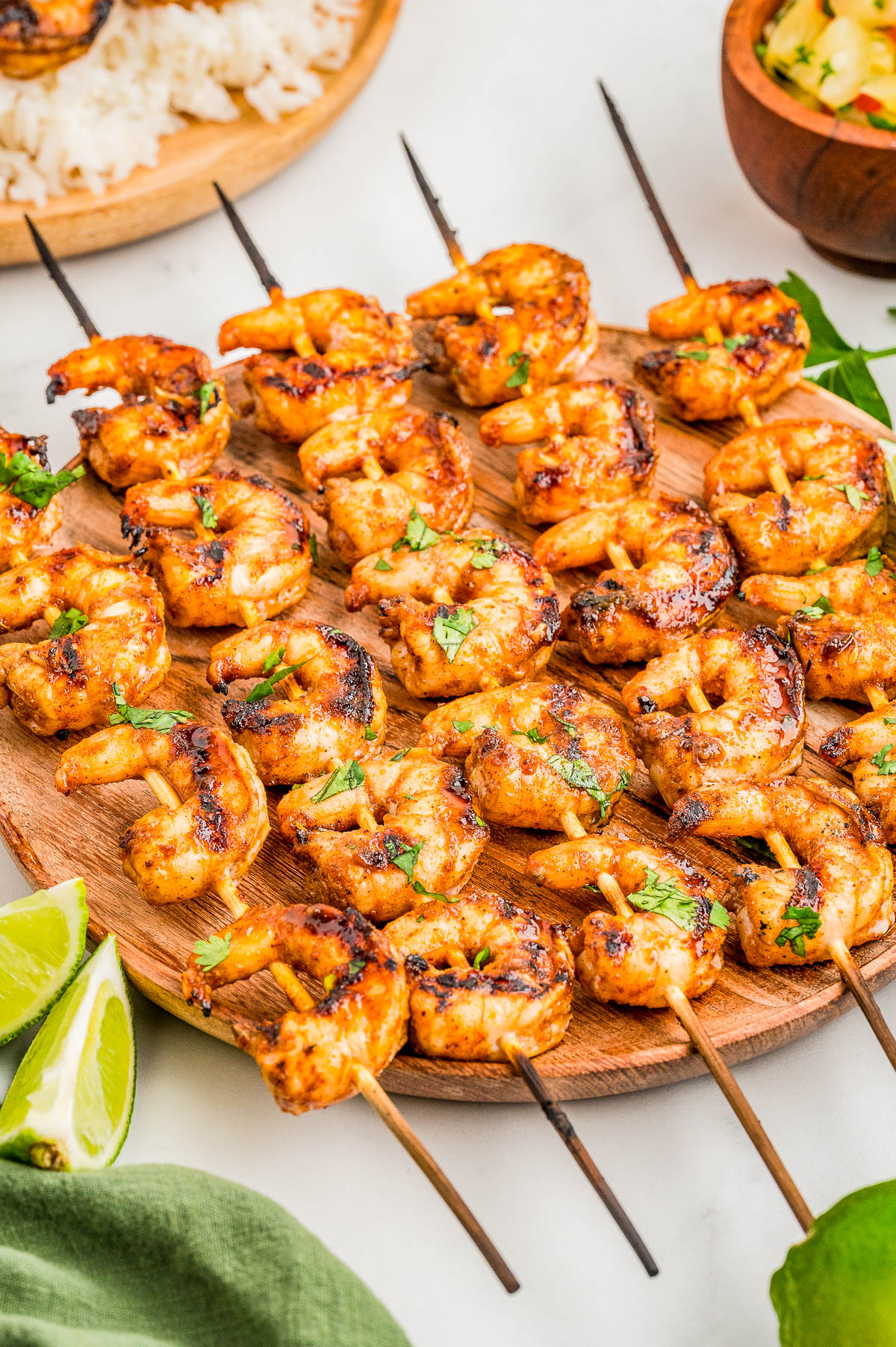 Grilled shrimp skewers served on a wooden plate, garnished with herbs, with lime wedges and other food items in the background.