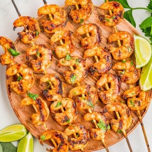 Grilled shrimp skewers arranged on a round wooden platter, garnished with chopped herbs and served with lime wedges.