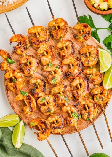 Grilled shrimp skewers arranged on a round wooden platter, garnished with chopped herbs and served with lime wedges.