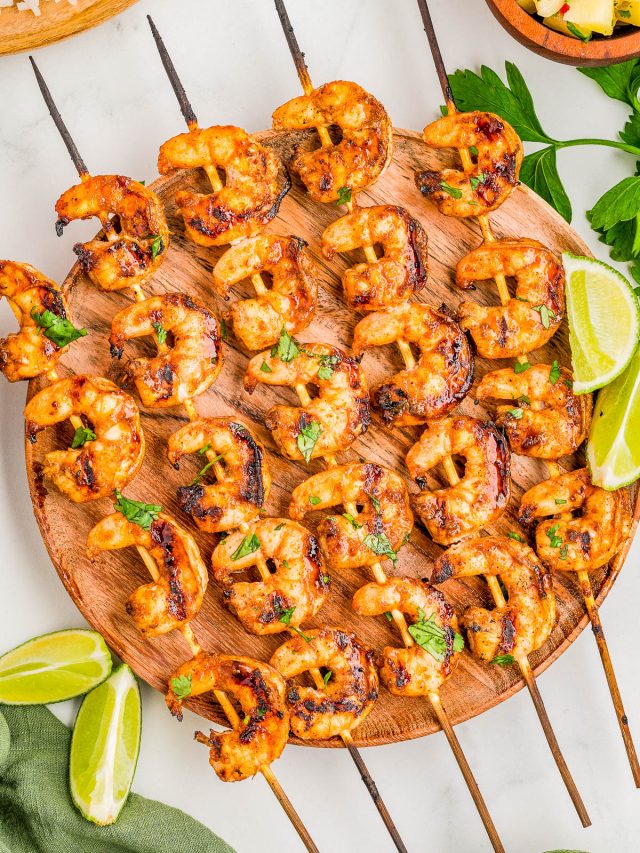 Grilled shrimp skewers arranged on a round wooden platter, garnished with chopped herbs and served with lime wedges.