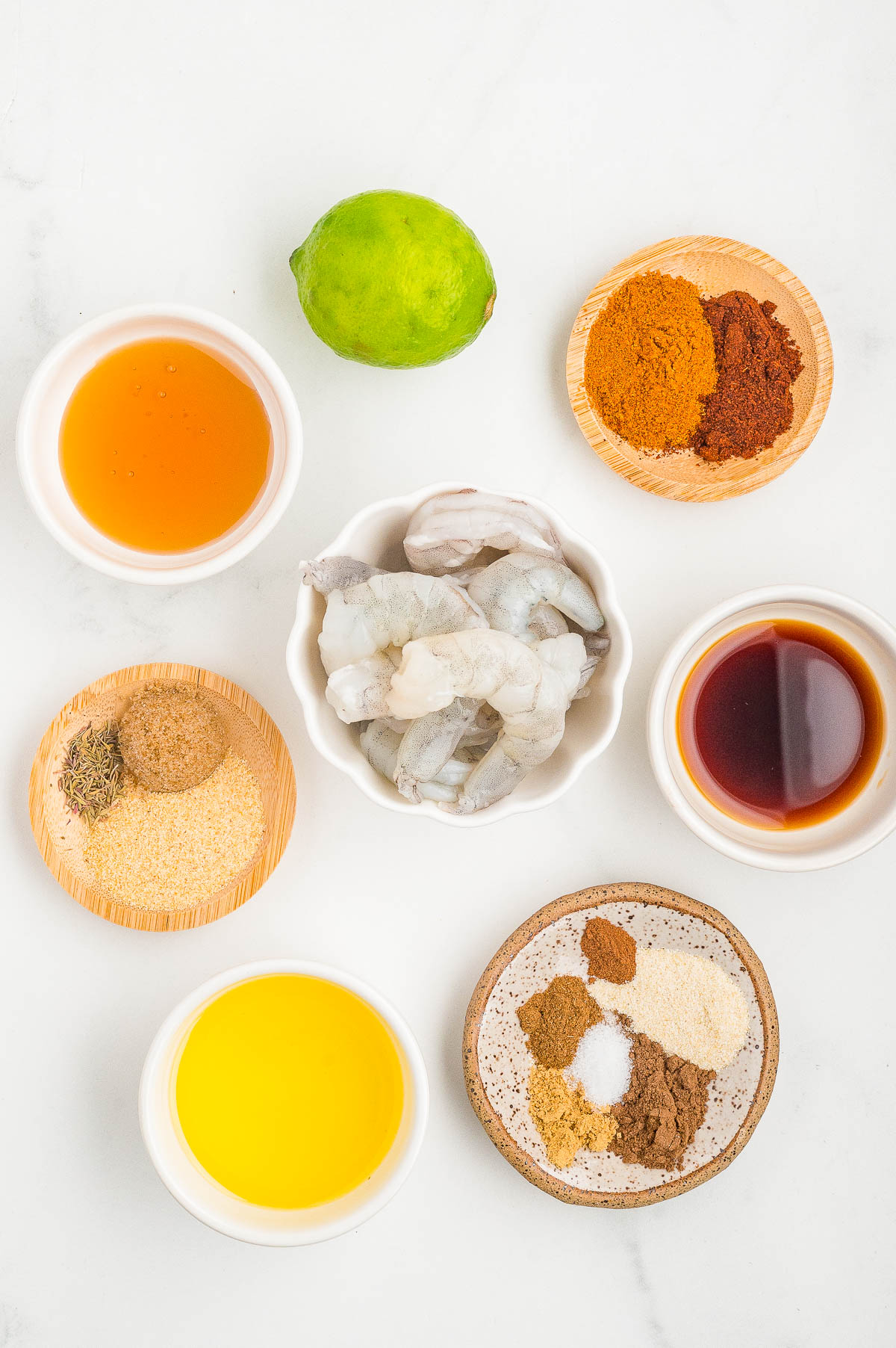 A variety of ingredients in small bowls, including peeled shrimp, spices, oil, lime, vinegar, and seasonings, arranged on a white surface.