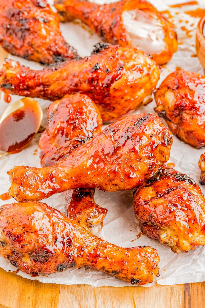 Several glazed and grilled chicken drumsticks are arranged on parchment paper, with sauce visible on the meat and a wooden surface beneath.
