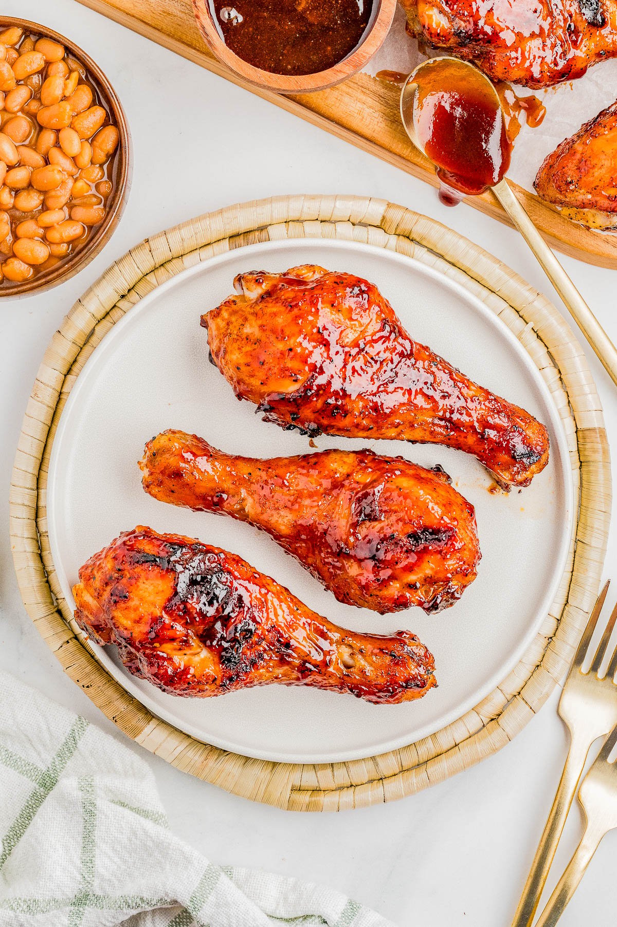 Three glazed barbecue chicken drumsticks on a white plate, with baked beans, sauce, and a spoon visible nearby.