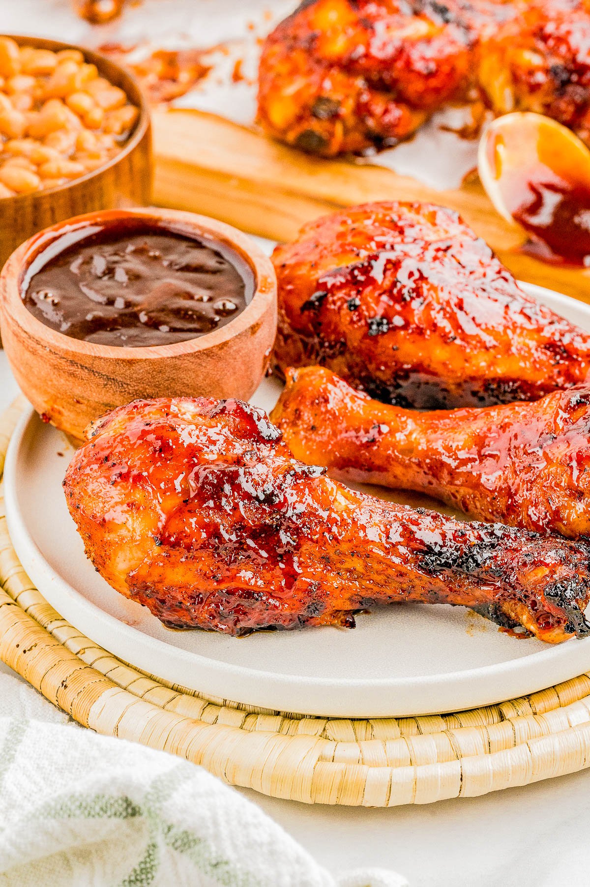 Grilled barbecue chicken drumsticks served on a white plate with a side of barbecue sauce and baked beans.