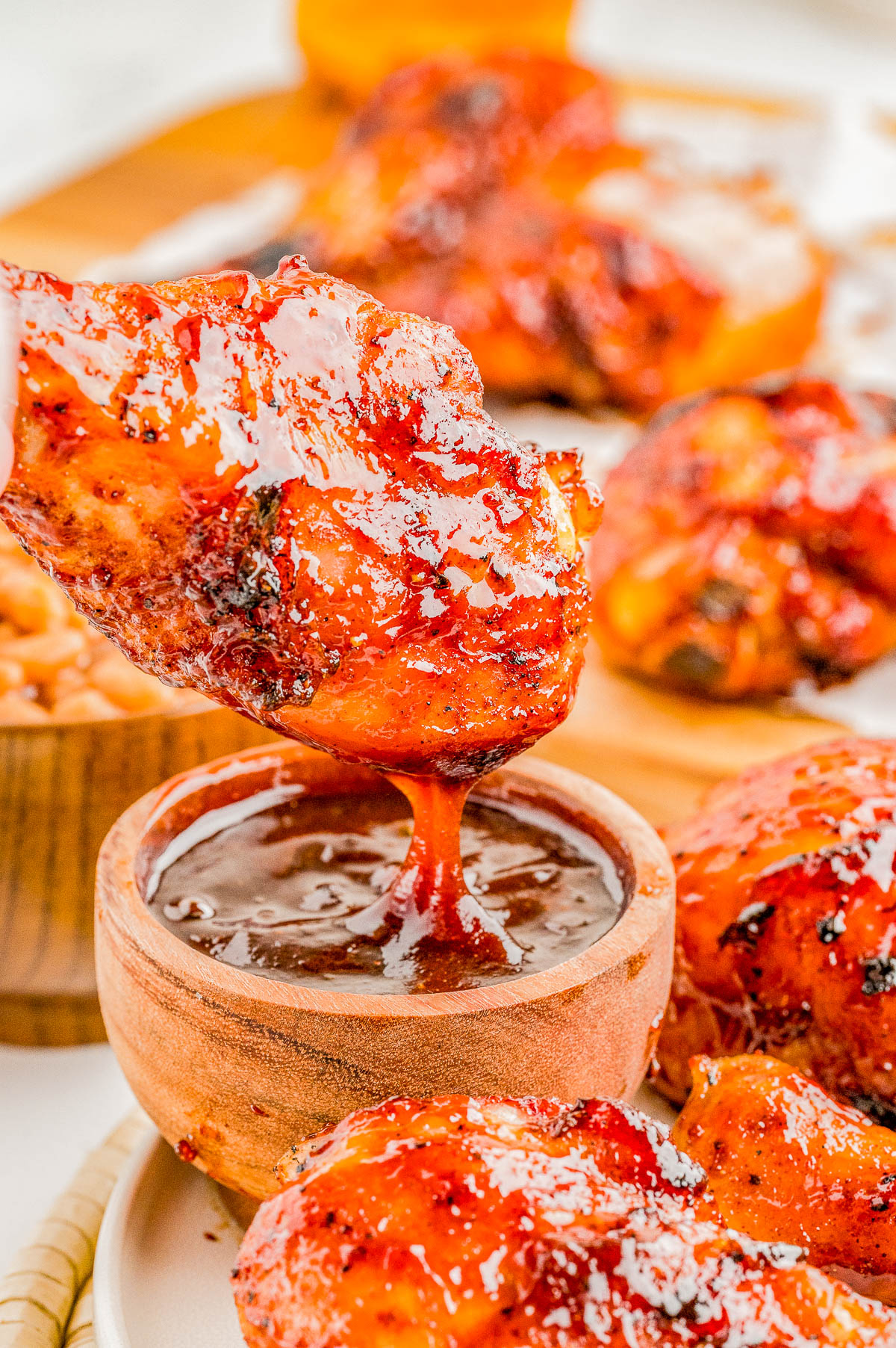 A piece of glazed grilled chicken is being dipped into a small wooden bowl of barbecue sauce, with more chicken pieces on a plate in the background.