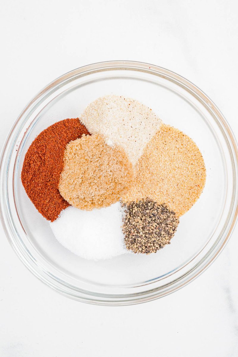 A glass bowl containing separate piles of assorted ground spices and seasonings on a white surface.