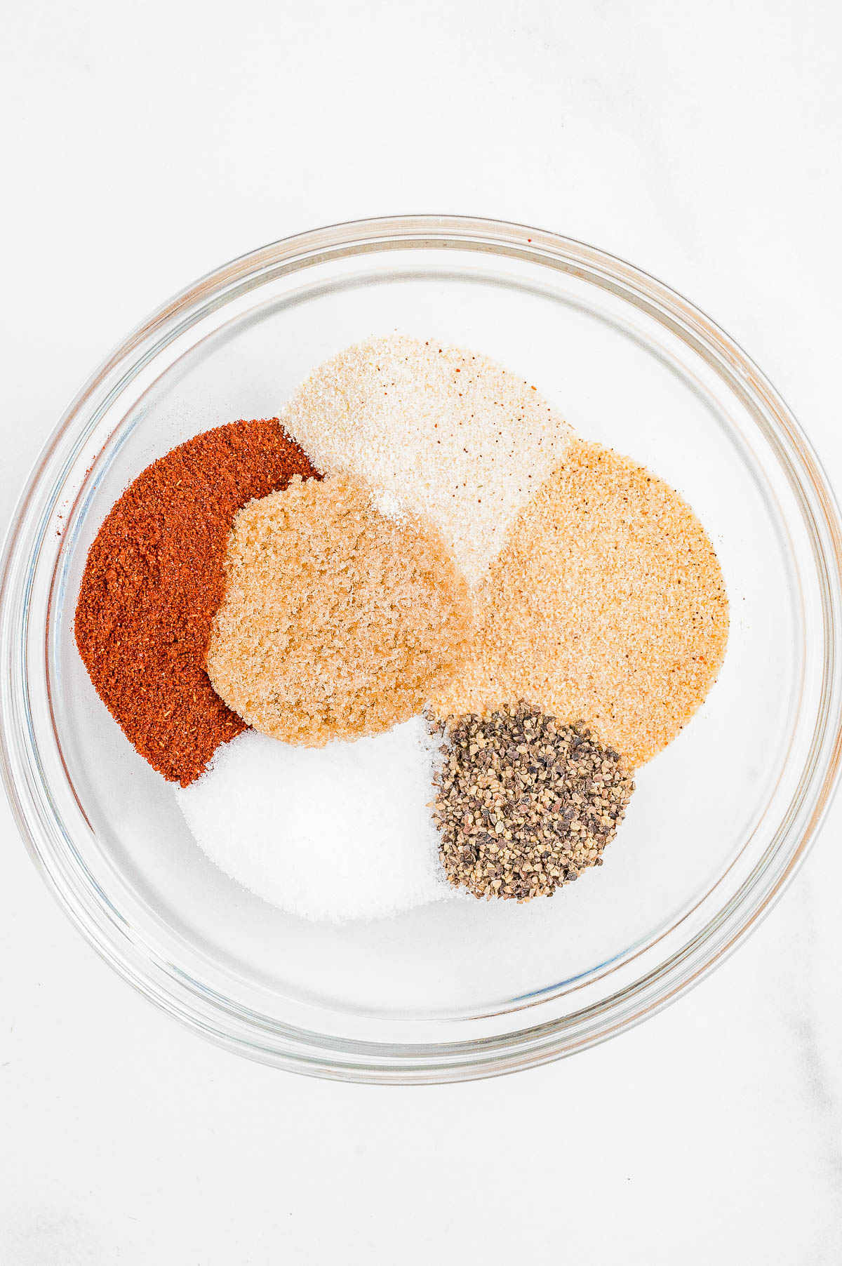 A glass bowl containing separate piles of assorted ground spices and seasonings on a white surface.