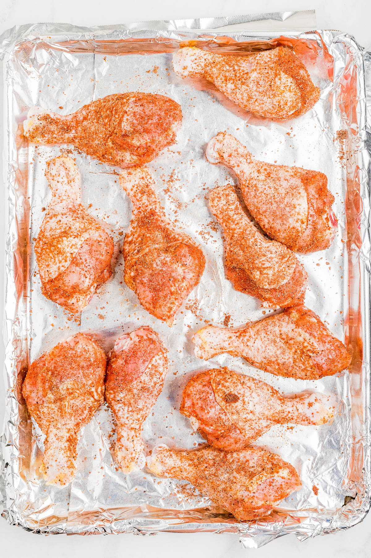 Raw chicken drumsticks coated with spices are arranged on a foil-lined baking sheet, ready for cooking.