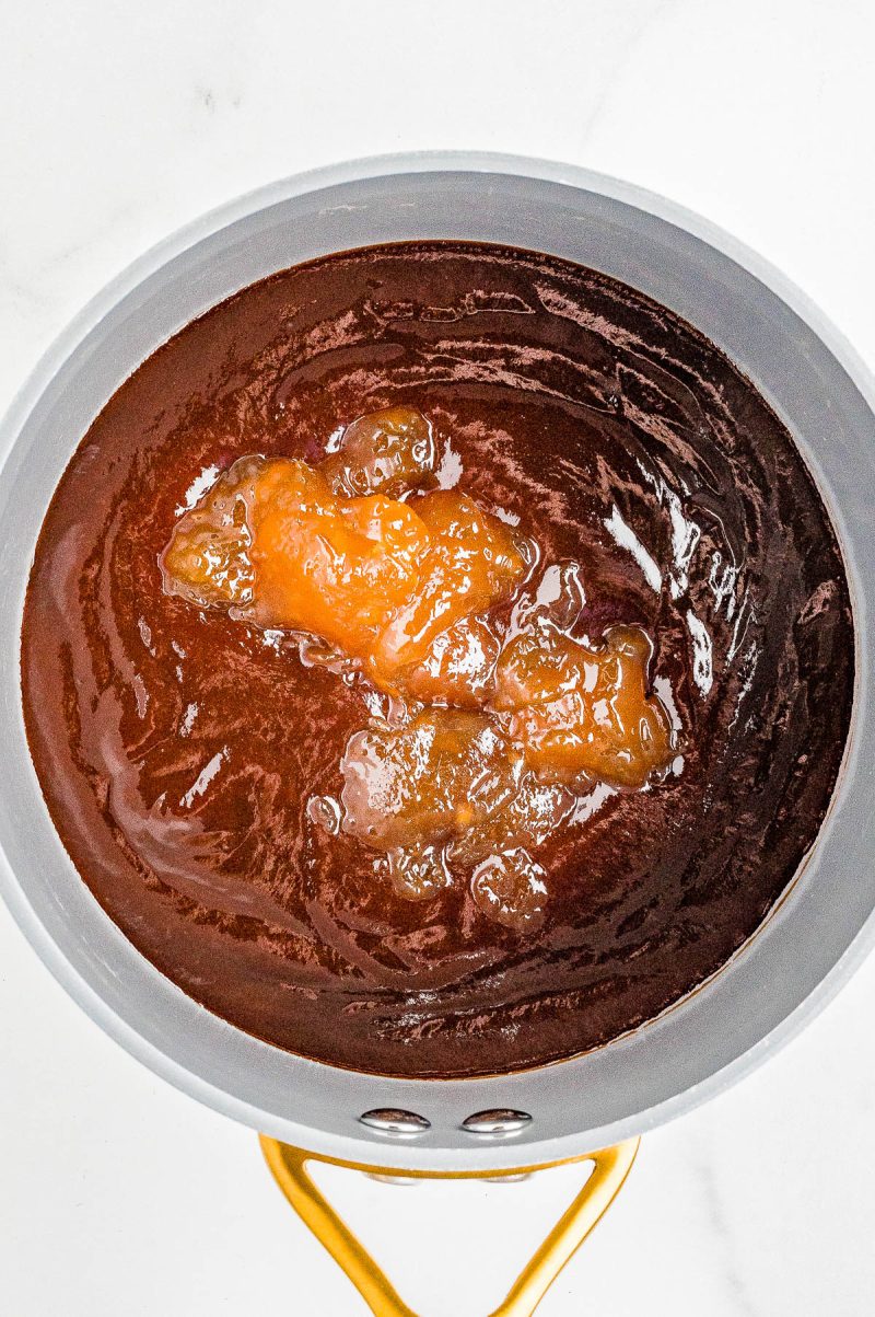 A saucepan filled with a dark red sauce and a dollop of yellowish jam or preserves on top, set on a white surface.