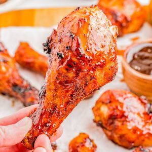 A hand holds a glazed, grilled chicken drumstick in front of a plate with more drumsticks and a bowl of barbecue sauce.