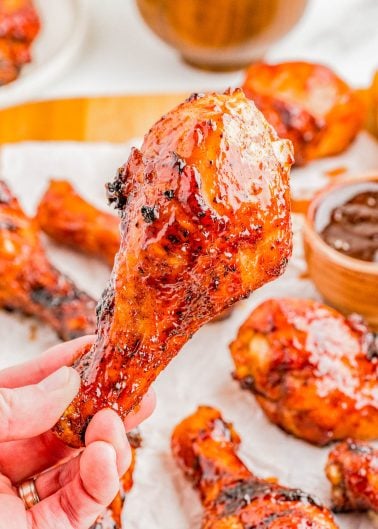 A hand holds a glazed, grilled chicken drumstick in front of a plate with more drumsticks and a bowl of barbecue sauce.