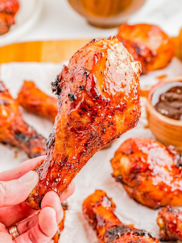 A hand holds a glazed, grilled chicken drumstick in front of a plate with more drumsticks and a bowl of barbecue sauce.