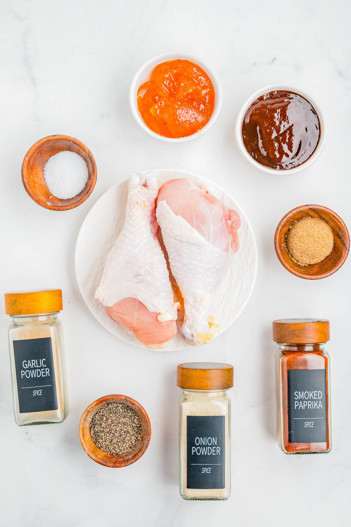 Raw chicken drumsticks on a plate surrounded by small bowls of salt, sugar, peach preserves, barbecue sauce, and jars of garlic powder, onion powder, smoked paprika, and black pepper.