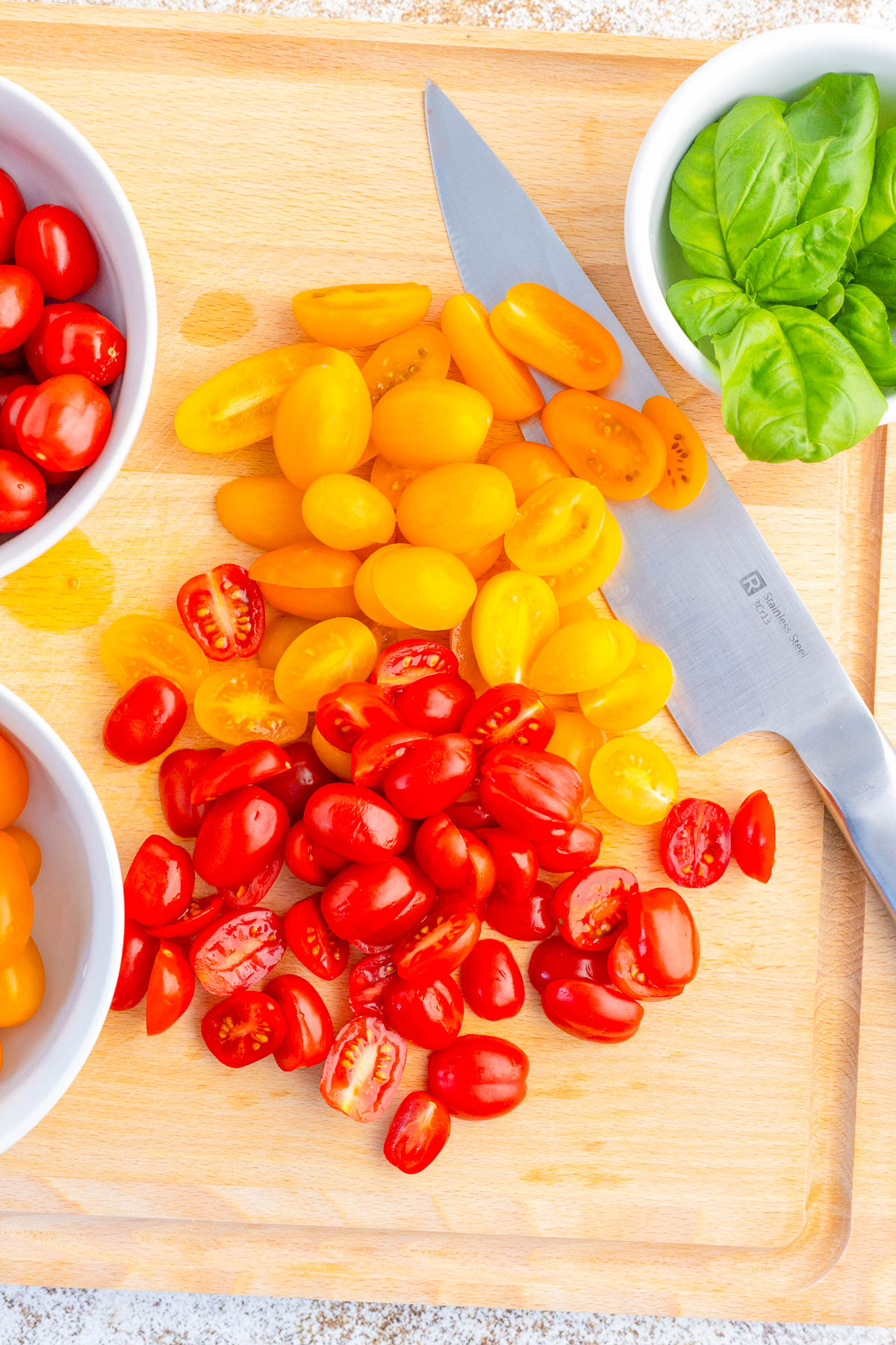 Sliced red and yellow cherry tomatoes on a wooden cutting board with a knife, two bowls of whole tomatoes, and a bowl of fresh basil leaves.