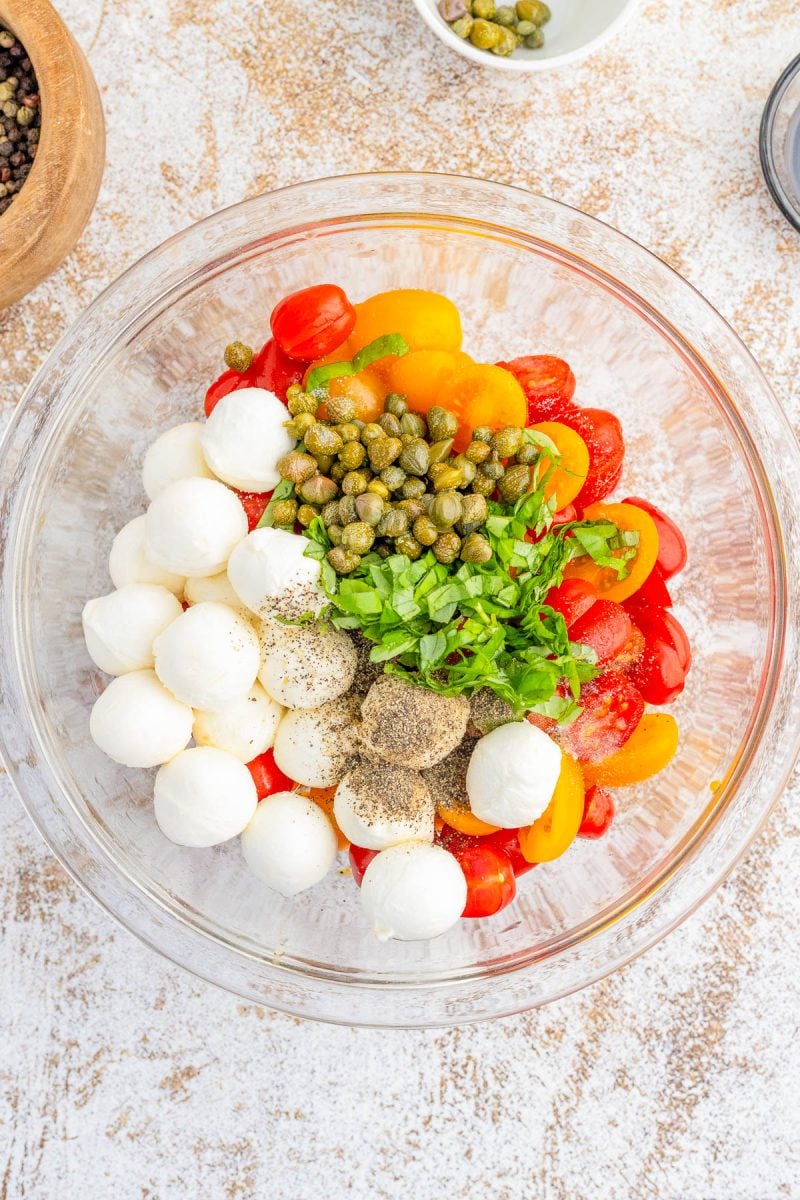 A glass bowl containing mozzarella balls, halved cherry tomatoes, capers, chopped parsley, and ground black pepper.