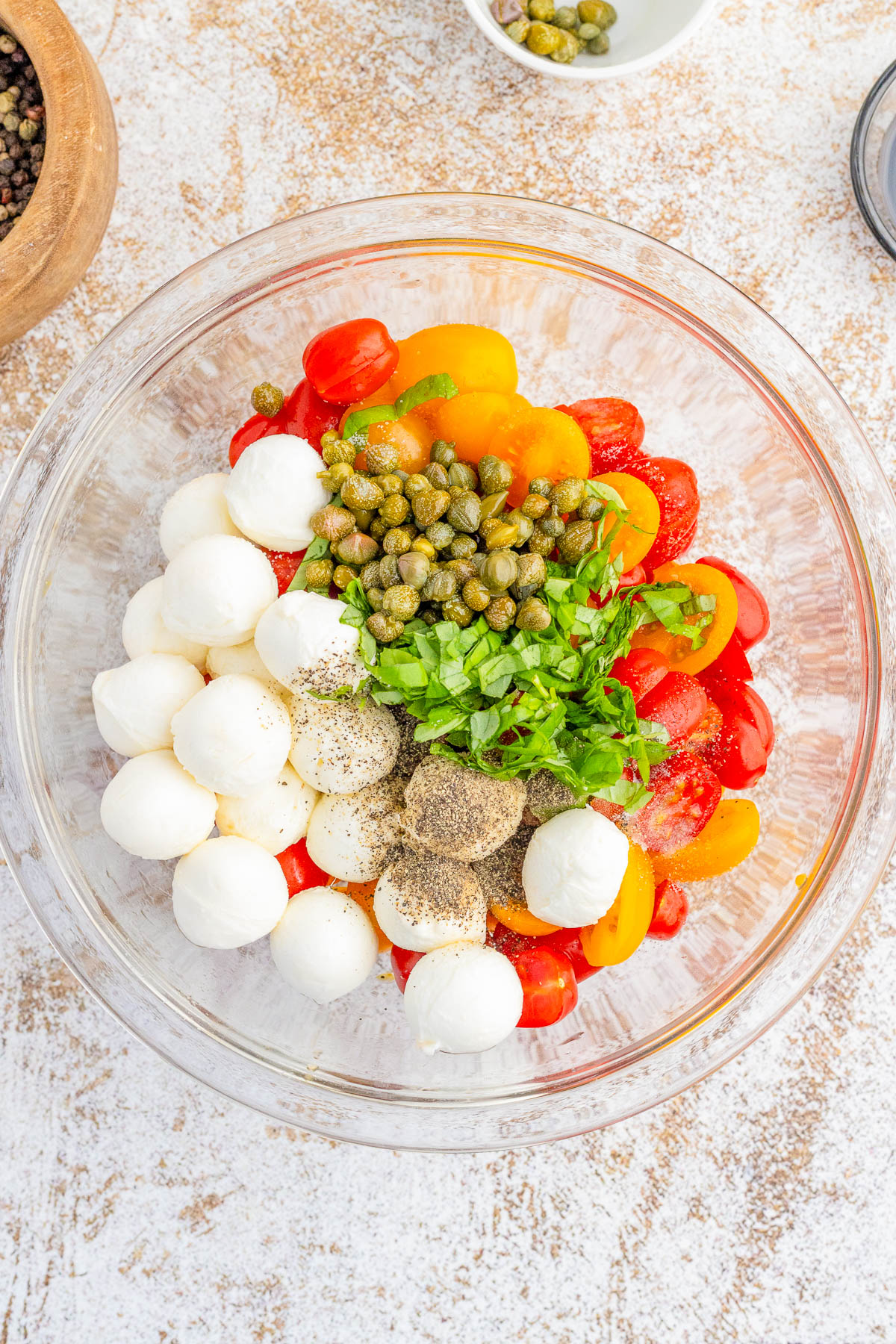 A glass bowl containing mozzarella balls, halved cherry tomatoes, capers, chopped parsley, and ground black pepper.