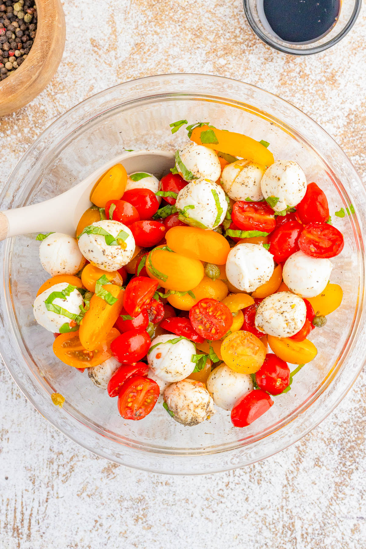 A glass bowl filled with cherry tomatoes, mozzarella balls, and fresh herbs, with a spoon for mixing. A bowl of pepper and a cup of balsamic vinegar are nearby.
