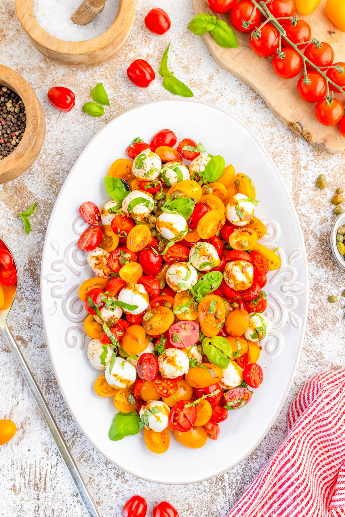 A white oval plate with a colorful salad of halved cherry tomatoes, mozzarella balls, and fresh basil, surrounded by ingredients on a light rustic surface.