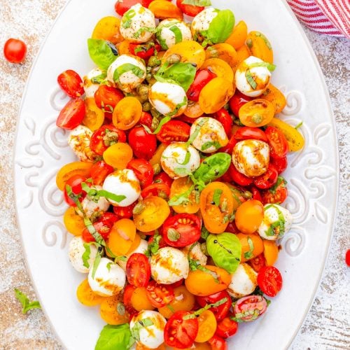 An oval white plate holds a salad of red and yellow cherry tomatoes, mozzarella balls, fresh basil leaves, and a light dressing.