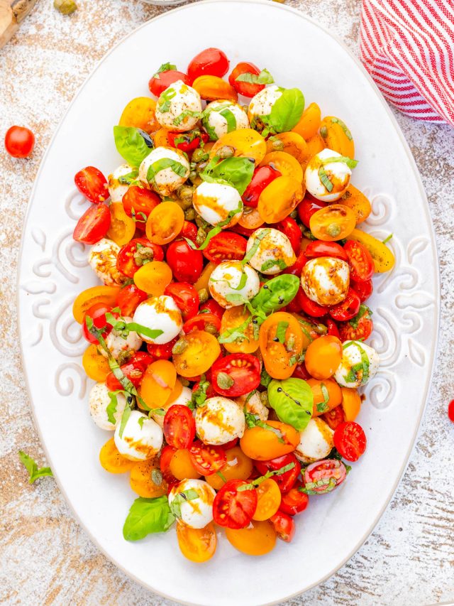 An oval white plate holds a salad of red and yellow cherry tomatoes, mozzarella balls, fresh basil leaves, and a light dressing.