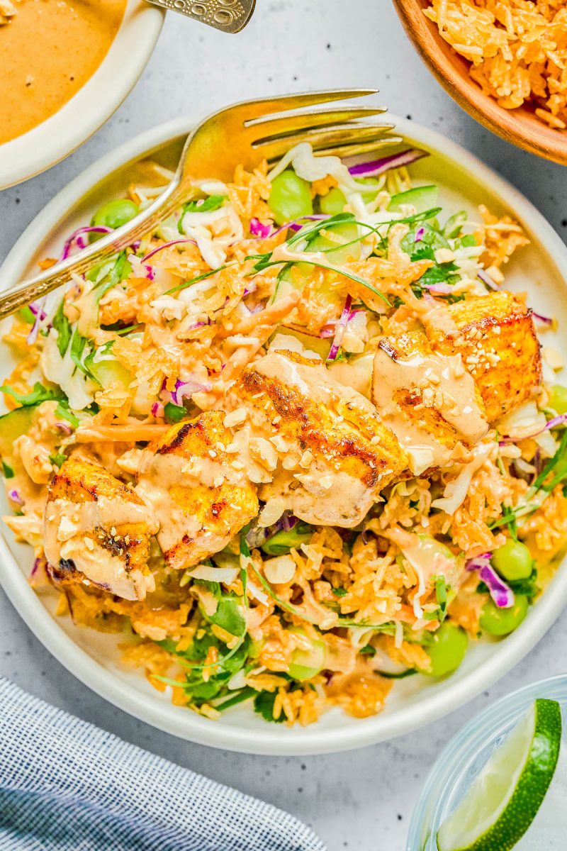 A plate of grilled chicken on a bed of rice, shredded cabbage, greens, and edamame, topped with sauce and crispy bits, with a fork and a lime wedge on the side.