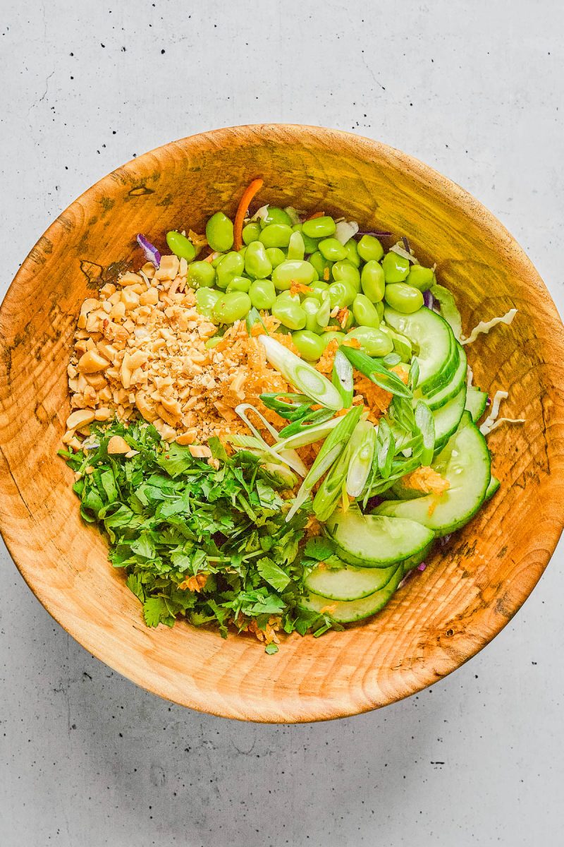 A wooden bowl containing sliced cucumbers, edamame, chopped peanuts, fresh cilantro, shredded carrots, and green onions on a light background.