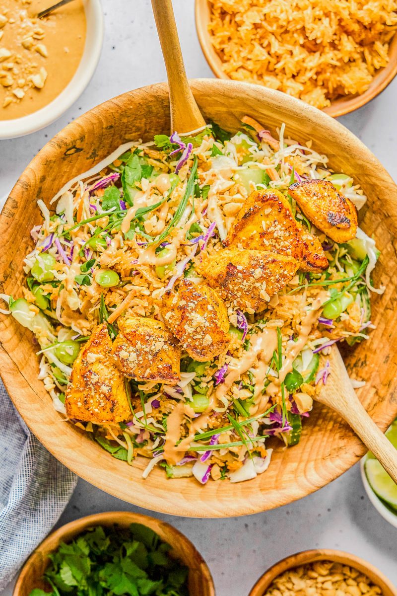 A wooden bowl filled with salad topped with grilled chicken pieces, shredded cabbage, greens, peanuts, and a creamy sauce, with bowls of rice, peanuts, and herbs nearby.
