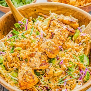 A wooden bowl filled with salad greens, shredded cabbage, grilled tofu, peanuts, and a creamy dressing, with wooden salad servers and side bowls of rice, cilantro, and lime wedges.