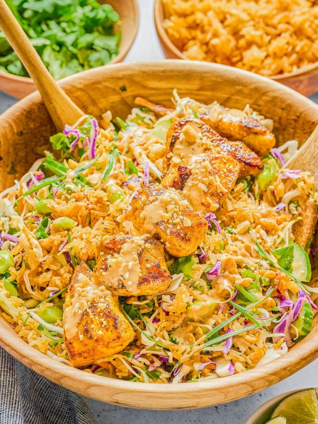 A wooden bowl filled with salad greens, shredded cabbage, grilled tofu, peanuts, and a creamy dressing, with wooden salad servers and side bowls of rice, cilantro, and lime wedges.