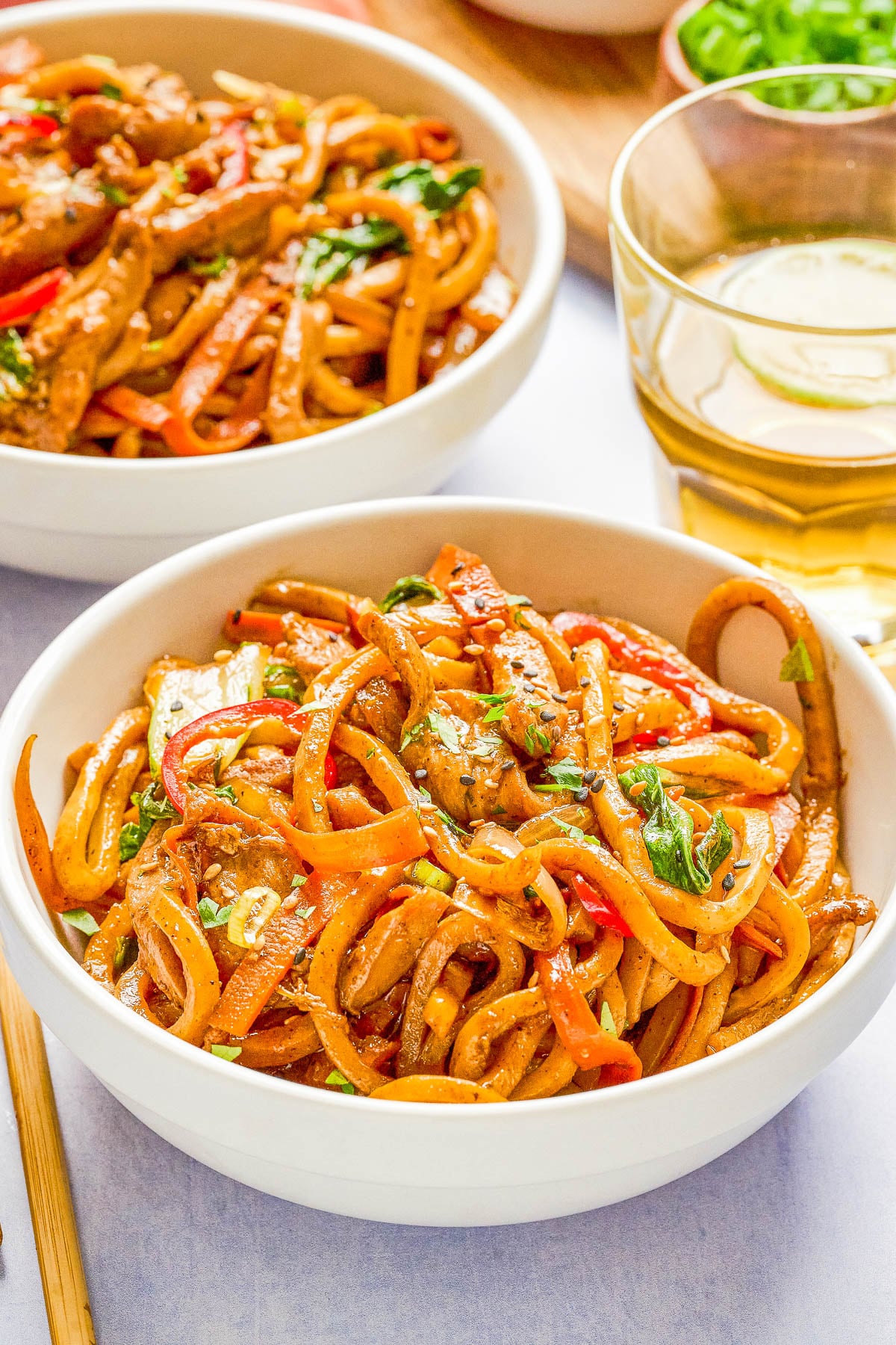 Two white bowls filled with stir-fried noodles, vegetables, and herbs are placed on a table next to a glass of iced beverage and a wooden chopstick.
