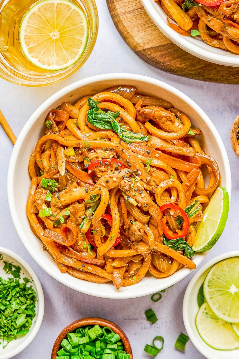 A bowl of stir-fried noodles with vegetables and sliced chicken, garnished with lime, sesame seeds, and green onions. Sliced lemons and herbs are visible nearby.