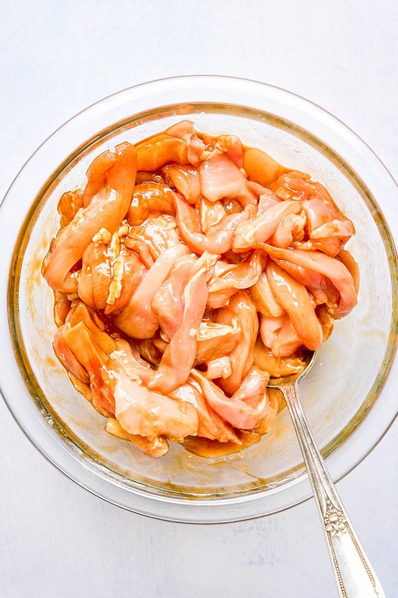 Raw chicken strips marinating in a glass bowl with a silver fork.