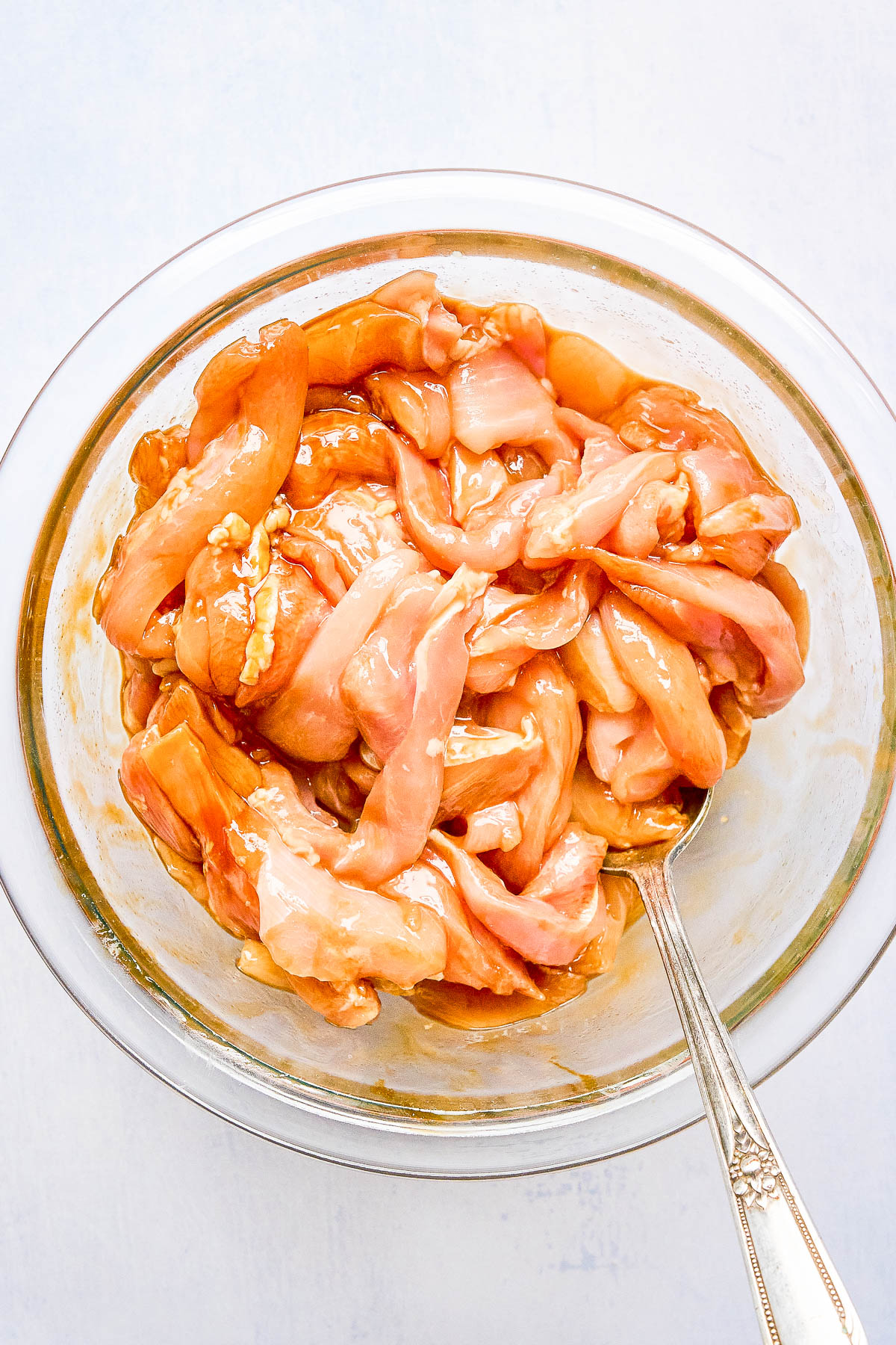 Raw chicken strips marinating in a glass bowl with a silver fork.