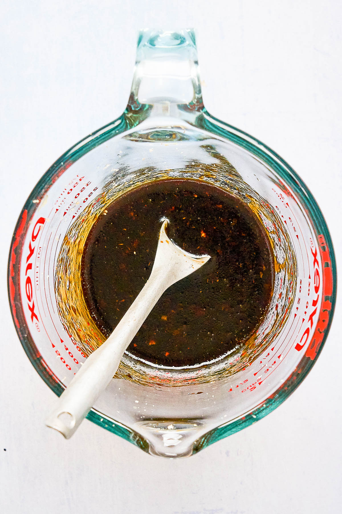 A glass measuring cup filled with a dark liquid sauce, stirred with a ceramic spoon, viewed from above.