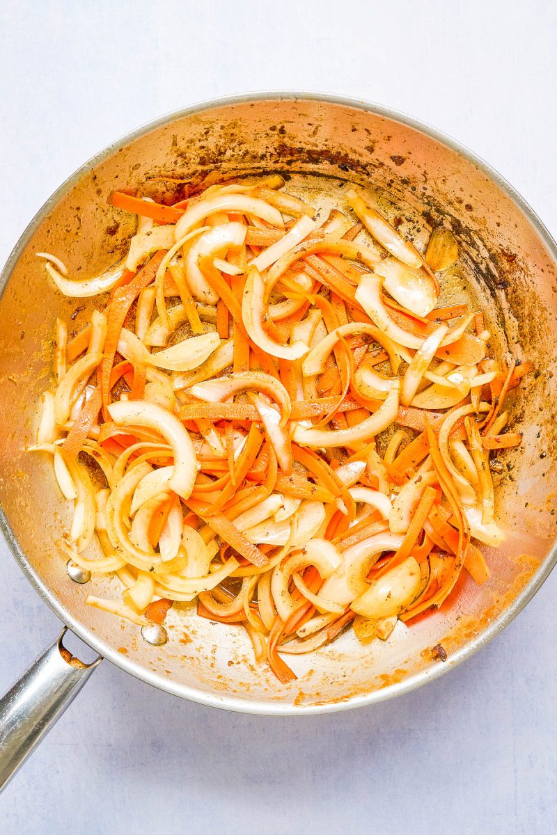 Sliced onions and carrots sautéing in a stainless steel pan with visible browning on the vegetables and pan.