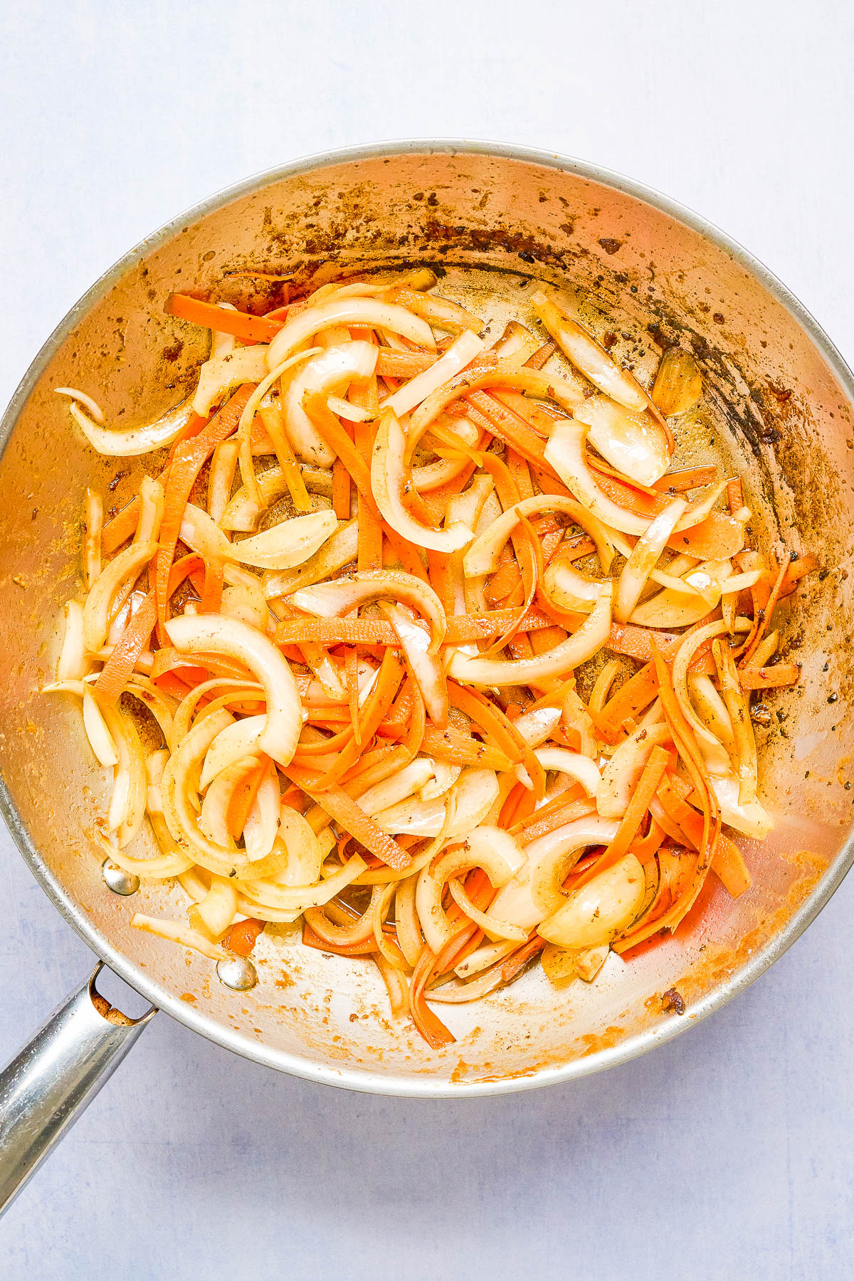 Sliced onions and carrots sautรฉing in a stainless steel pan with visible browning on the vegetables and pan.