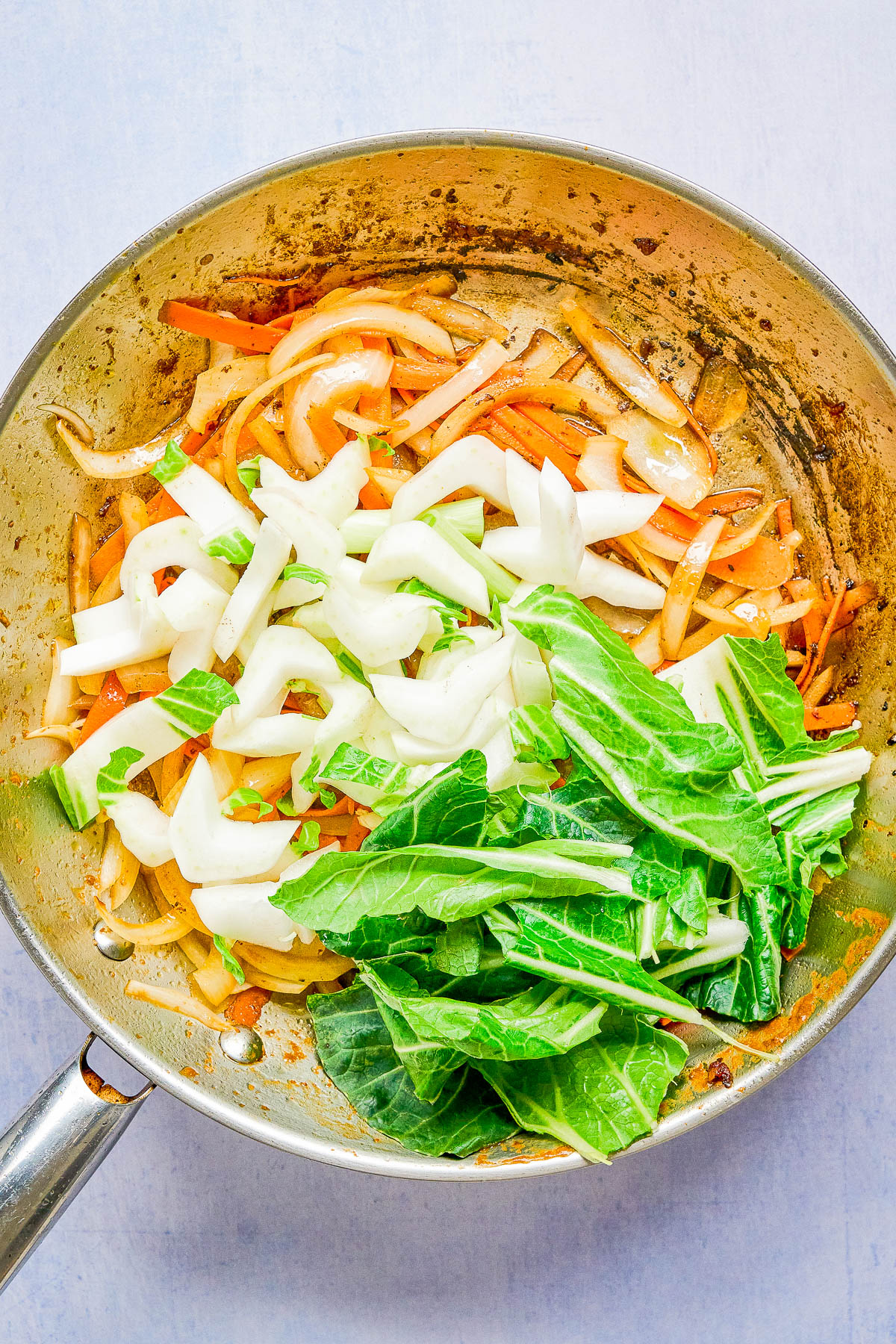 A stainless steel pan with sliced onions, orange bell peppers, and fresh chopped bok choy being sautรฉed.