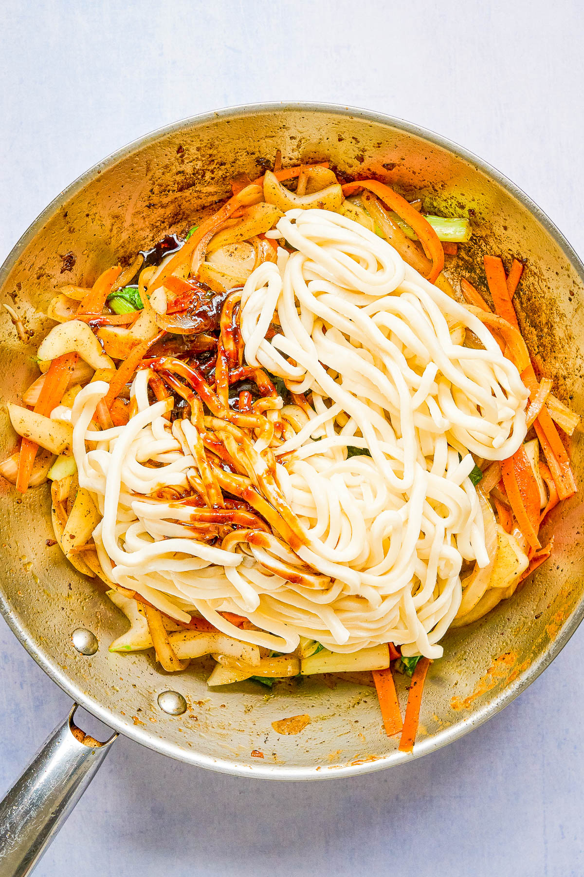 A pan with cooked udon noodles, sliced carrots, onions, and green vegetables, topped with a dark sauce, ready to be mixed.
