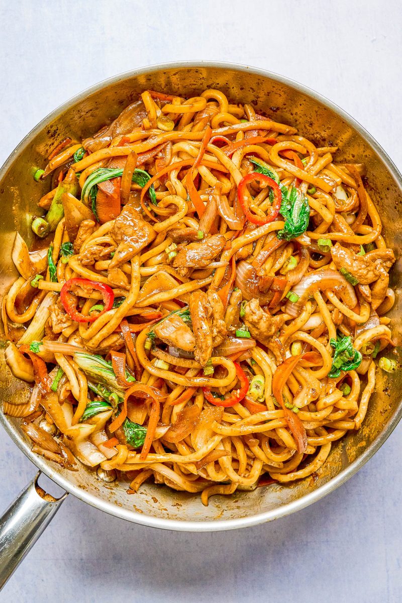 A skillet filled with stir-fried noodles, sliced beef, bell peppers, bok choy, onions, and a savory sauce.