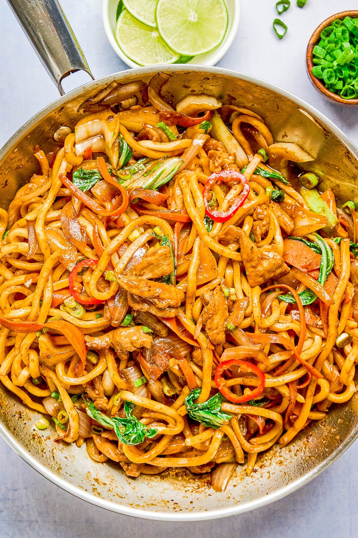 A pan filled with stir-fried noodles, vegetables, and sliced meat, garnished with red chili and green onion, with lime wedges and scallions in small bowls nearby.