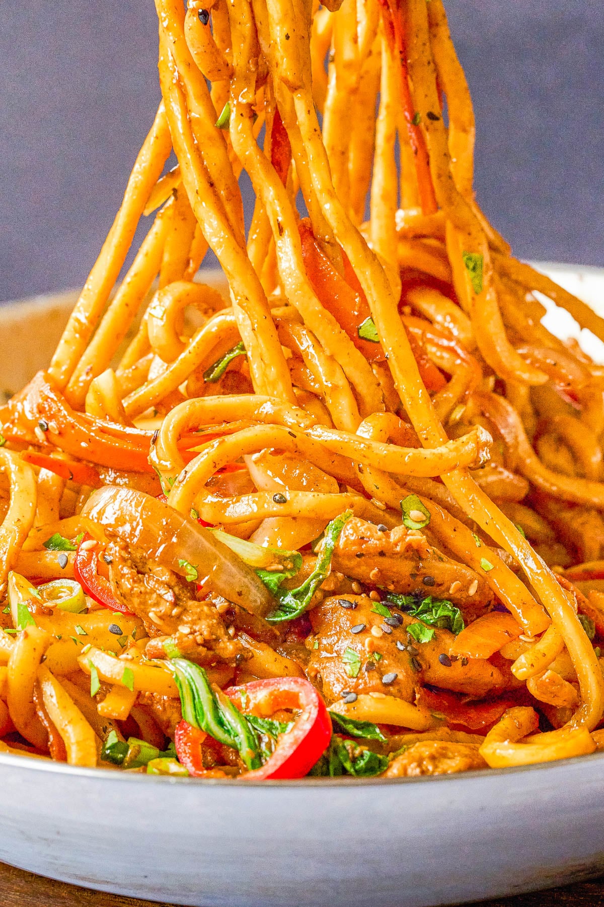 A close-up of a bowl of stir-fried noodles with vegetables, sliced meat, and herbs, with chopsticks lifting a portion from the bowl.