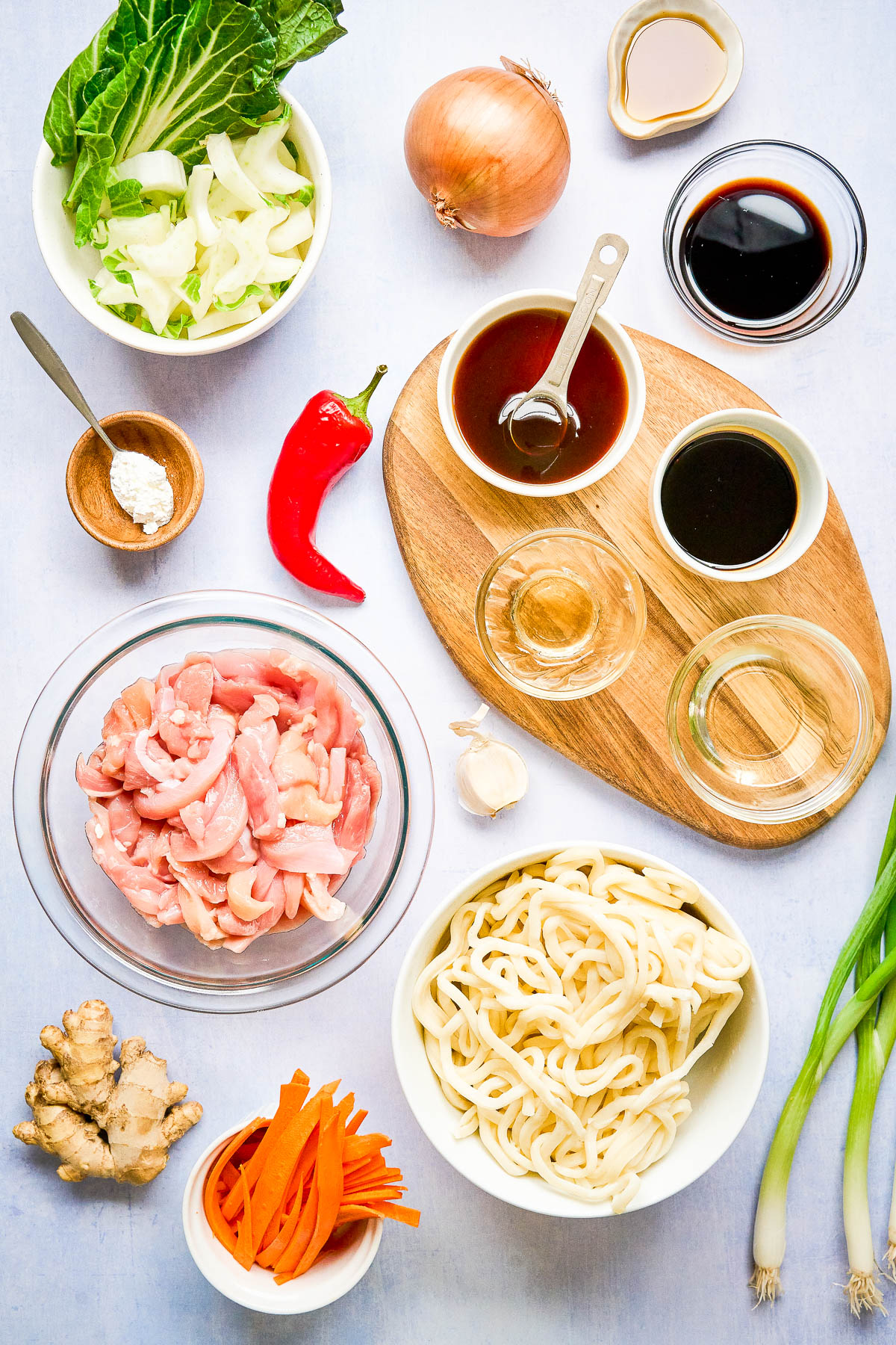 Various ingredients for a stir fry are arranged on a light surface, including sliced chicken, udon noodles, sauces, vegetables, fresh ginger, and a red chili pepper.