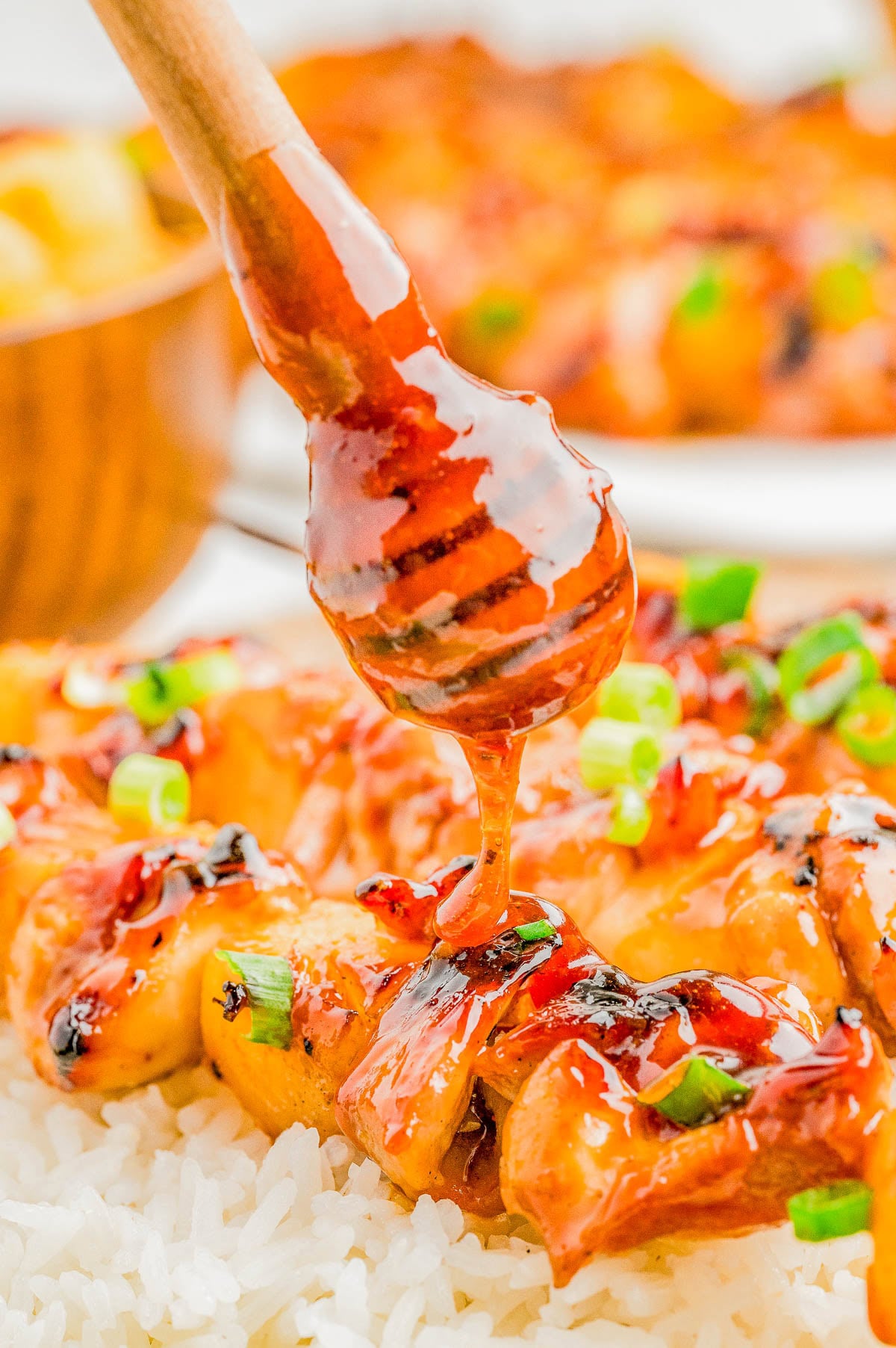 A honey dipper drizzles glaze over skewered grilled chicken served on a bed of white rice, garnished with chopped green onions.