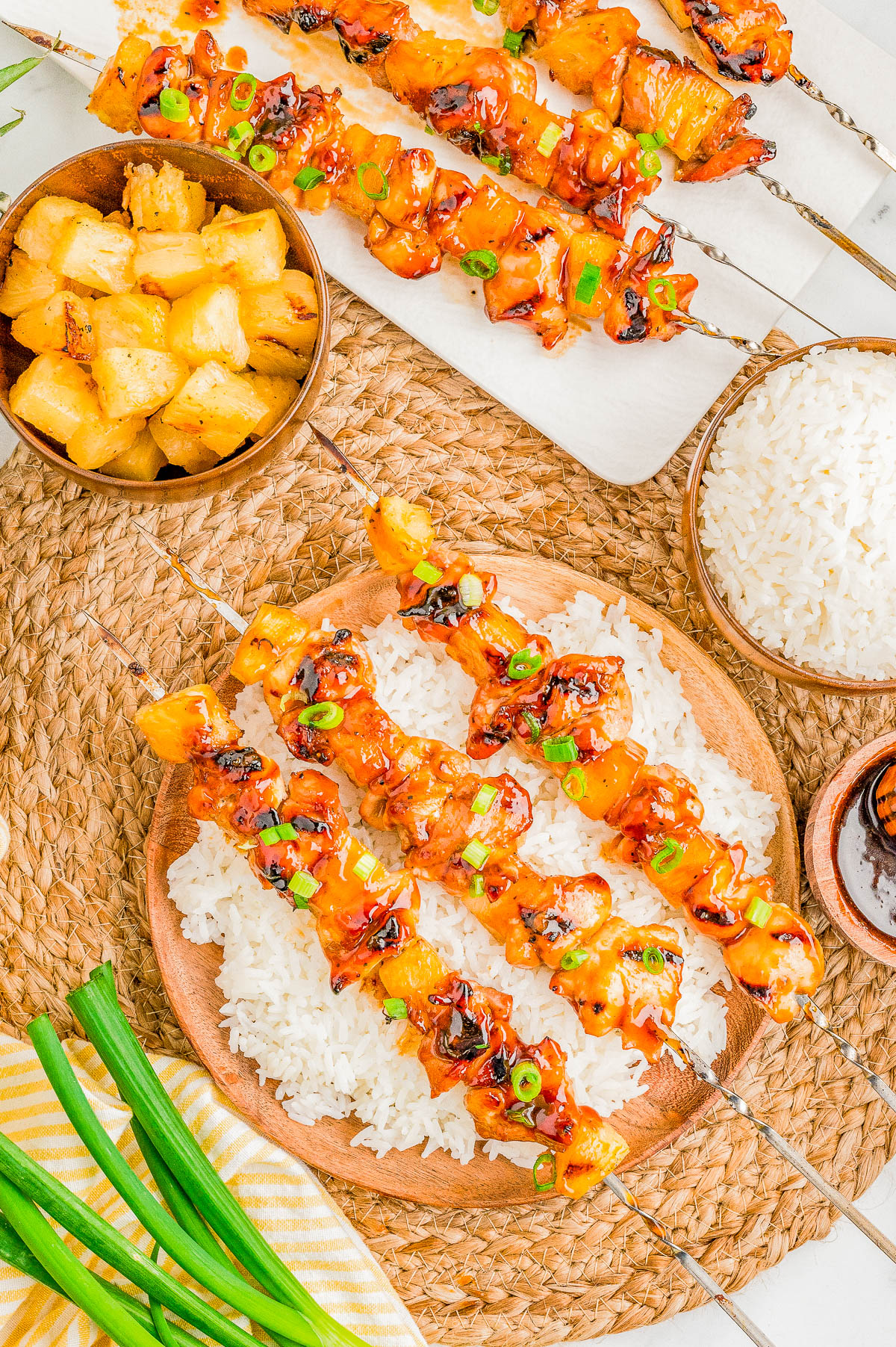Grilled chicken and pineapple skewers served on white rice, with bowls of pineapple chunks and rice on a woven placemat, and a bunch of green onions nearby.