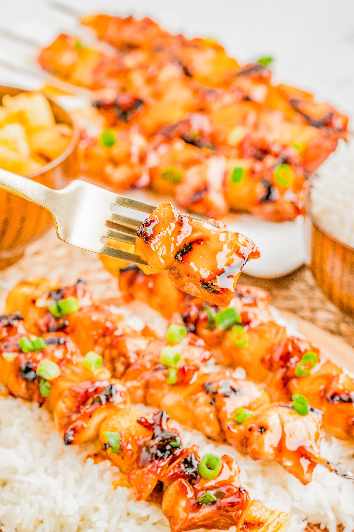 A fork holds a piece of glazed grilled chicken skewer garnished with green onions, with more skewers on rice and bowls of pineapple and rice in the background.